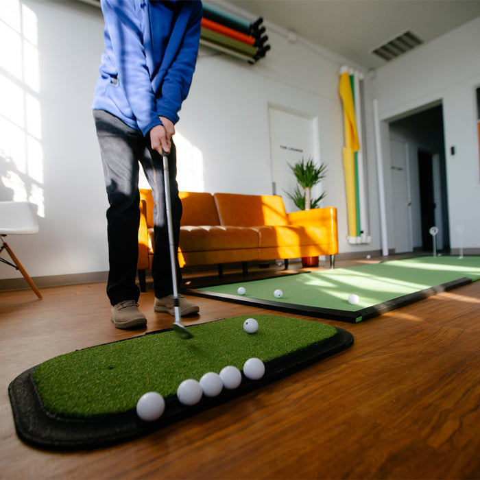 Someone practices putting with golf balls on a Birdie Turf indoor golf practice mat by BirdieBall in a room featuring a sofa, plant, and colorful wall tubes.