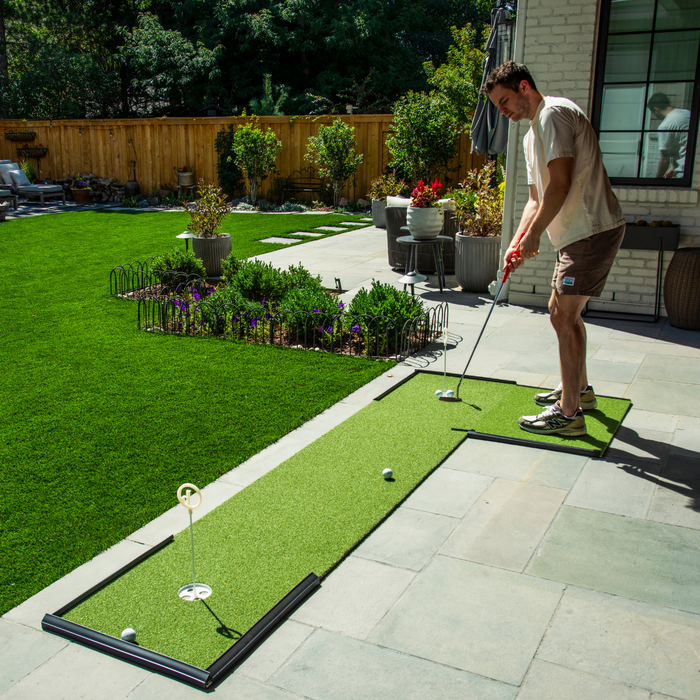 A man practices putting on a BirdieBall Golf Stance Mat (Options Available), set up as a putting green on a stone patio in his backyard garden.