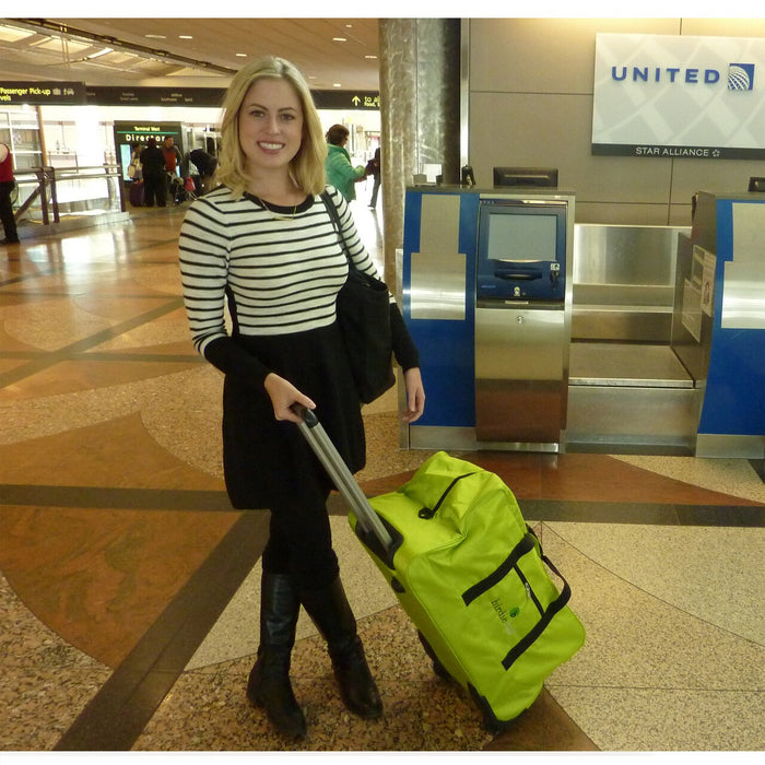 A woman stands in an airport near a United check-in kiosk, holding the handle of a bright green BirdieBall Wheeled Duffle Bag.