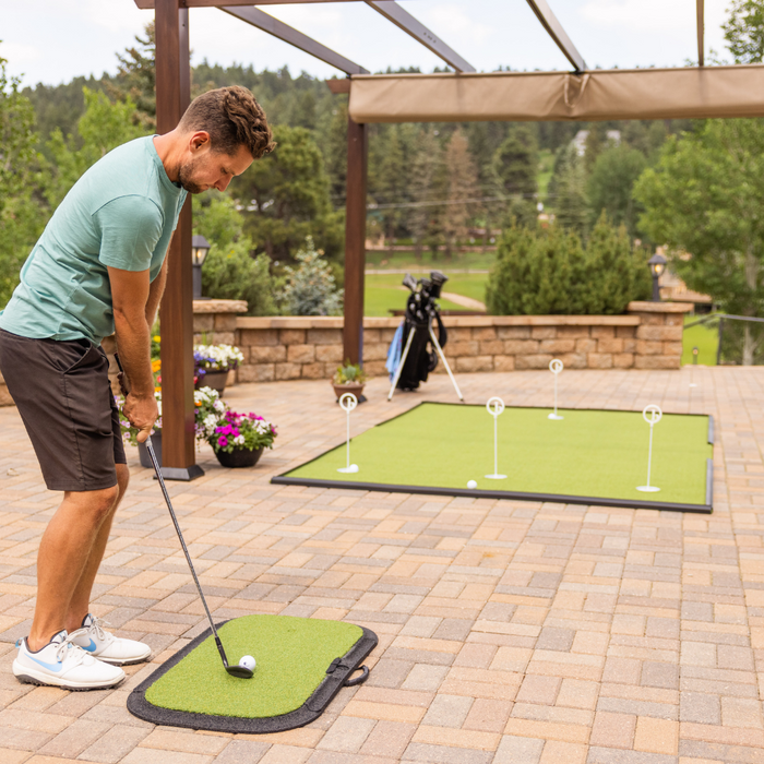 A man in a green shirt practices putting on the BirdieBall Birdie Turf golf mat on a patio, with golf flags and a golf bag visible in the background.