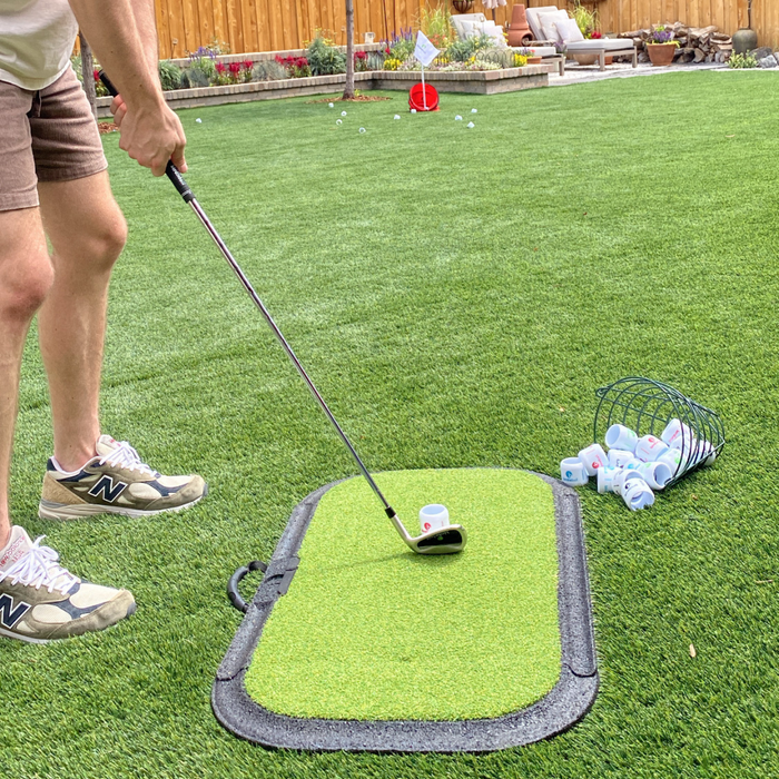 A person practices golf putting on the Birdie Turf by BirdieBall, standing next to a basket of golf balls and a small putting mat set up on artificial turf.