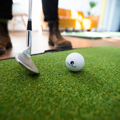 A close-up of the BirdieBall Birdie Turf chipping mat with a golf club next to a ball, and a person in boots standing nearby indoors.