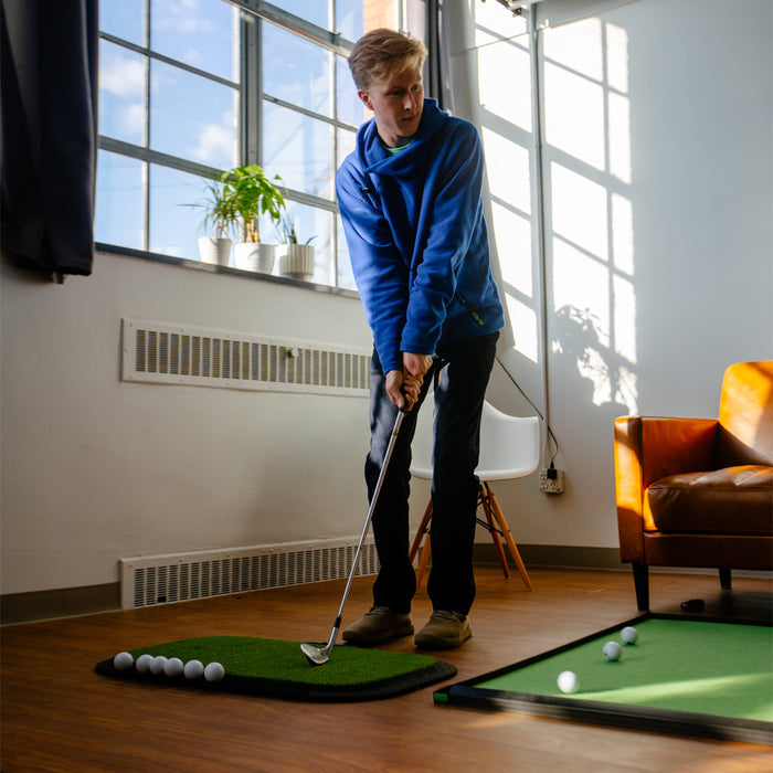 A person in a blue hoodie practices putting on the Birdie Turf by BirdieBall in a bright room with wooden floors and modern furniture.