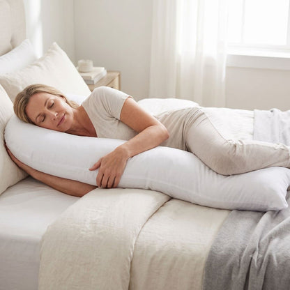 A woman sleeping peacefully on a bed using a white j-shaped body pillow, dressed in light beige loungewear. The bedroom is softly lit with natural light from a window, featuring neutral-toned bedding and a bedside table.