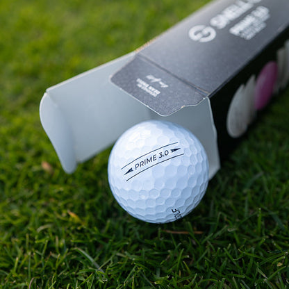 A BirdieBall Dozen Golf Balls (Snell Golf PRIME 3.0) with a urethane cover, featuring a white ball partially out of an open black box, is shown resting on green grass.