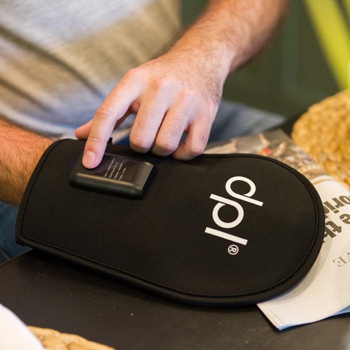 A close-up of a person's hand pressing a small black button on a black red light therapy hand mitt with a white 'pdl' logo, set on a dark tabletop.