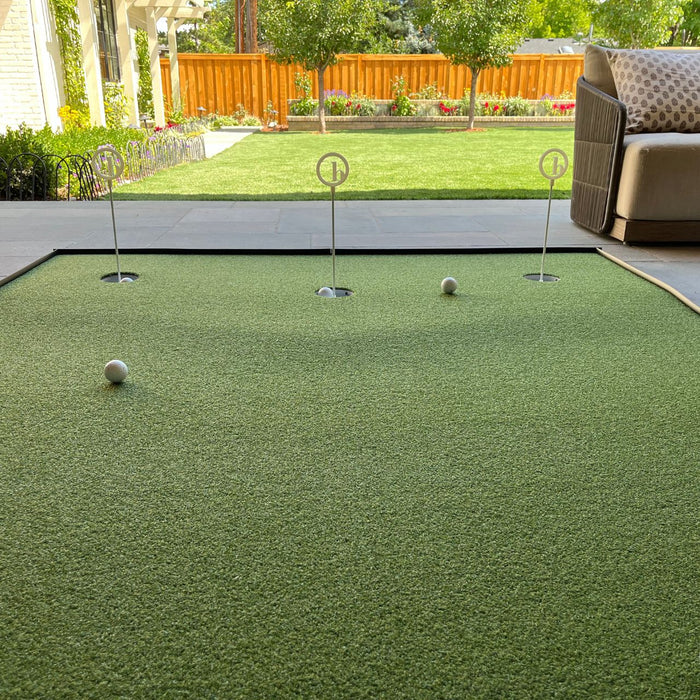 A BirdieBall Full Width Hole Deepening/Break Shim (for use with turf mats) is set up on a patio putting green, featuring tour-grade turf, three holes, and two golf balls, overlooking a grassy backyard with trees and a wooden fence.