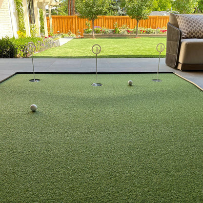 A BirdieBall Full Width Hole Deepening/Break Shim (for use with turf mats) is set up on a patio putting green, featuring tour-grade turf, three holes, and two golf balls, overlooking a grassy backyard with trees and a wooden fence.