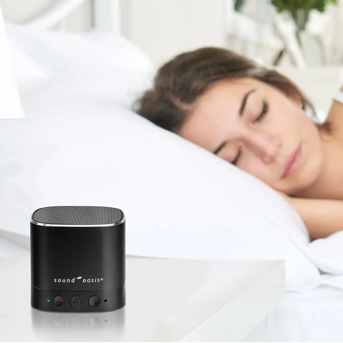 A woman lies in bed with her eyes shut, while the Sound Oasis sits on the bedside table.
