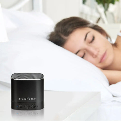A woman lies in bed with her eyes shut, while the Sound Oasis sits on the bedside table.