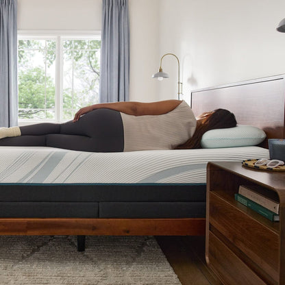 Person lying on a mattress in a bedroom with wooden furniture and large windows.