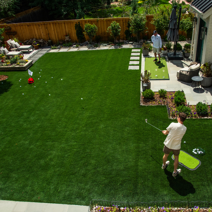 Two people play The Original Backyard Golf Game by BirdieBall on a lush lawn, aiming for golf targets using BirdieBall golf balls with a flag surrounded by garden landscaping.