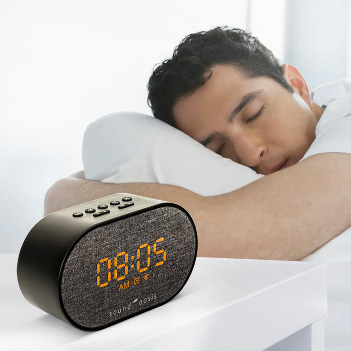 A man sleeping soundly in bed next to a black digital alarm clock with a textured face displaying 00:05 in orange digits.