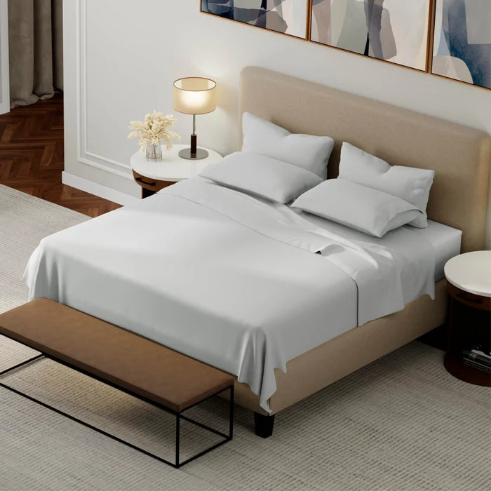 A neatly made bed with grey bedding and pillows, set in a modern bedroom with a beige upholstered headboard, bedside tables, a lamp, and a bench at the foot of the bed.